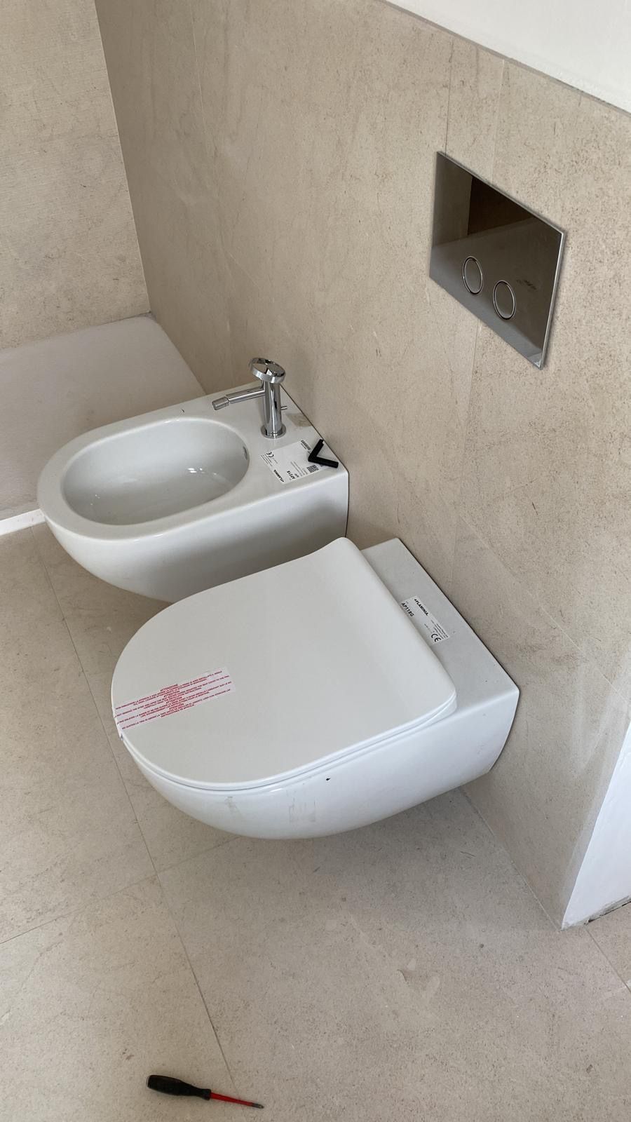 Piccolo bagno beige con WC sospeso, bidet e scopino con manico nero sul pavimento.