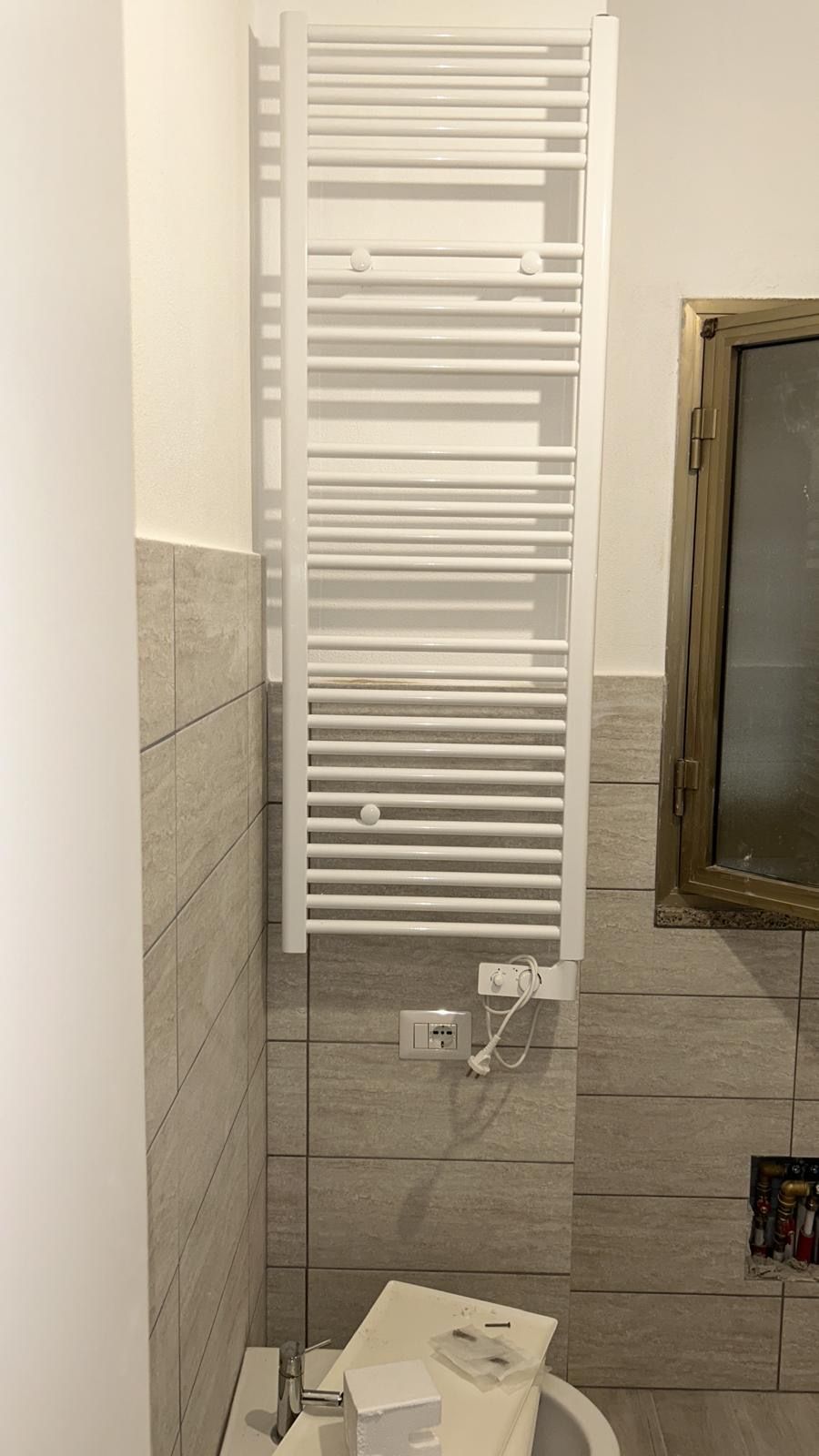 Scaldasalviette bianco a parete in un bagno piastrellato con lavabo e specchio sulla destra.