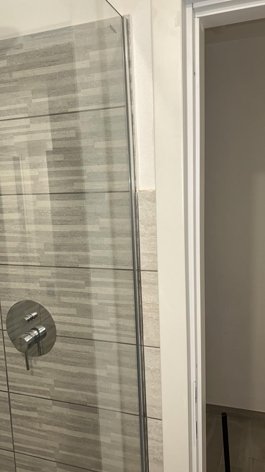 Doccia con porta in vetro e parete piastrellata accanto alla porta aperta del bagno.