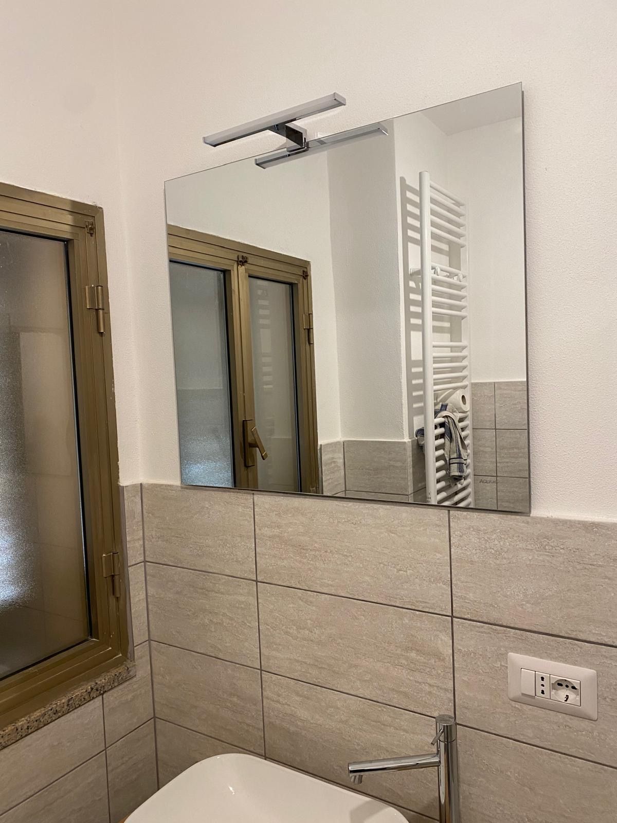 Specchio da bagno sopra il lavabo piastrellato di beige con riflesso della finestra e applique a parete.