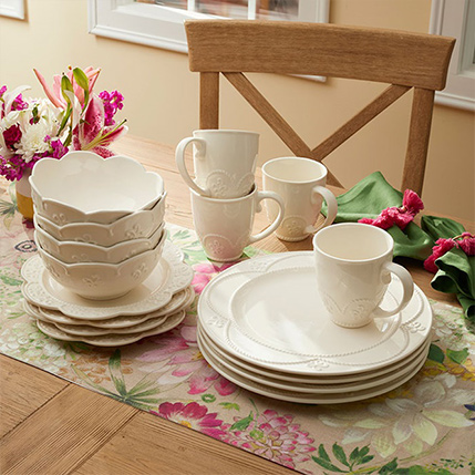 Stacked white dishes and teacups on a floral table setting with flowers and a wooden chair in the background