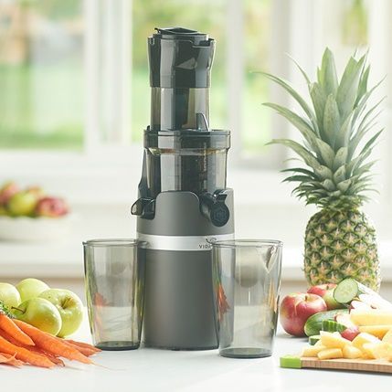 Juicer with pineapple, apples, carrots, and empty glasses on a bright kitchen counter
