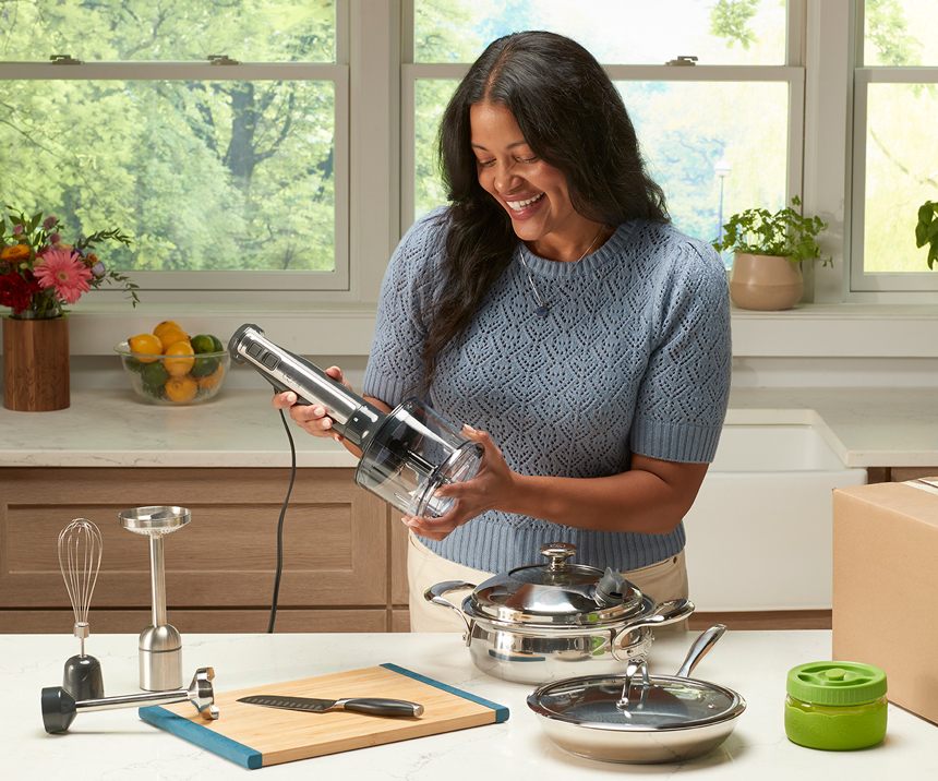 Woman in kitchen holding a hand mixer, smiling beside cookware and utensils on the counter.