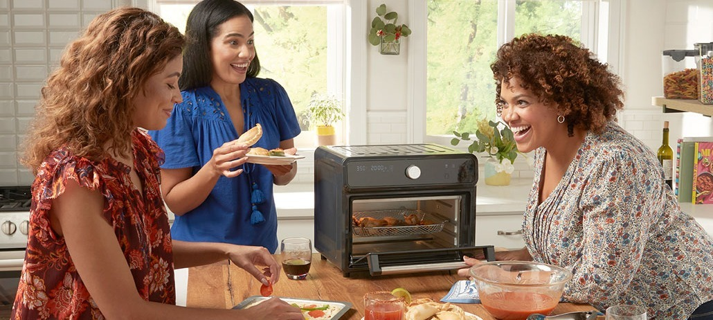 Three people chatting around a small table beside a lit tabletop fireplace in a bright living room