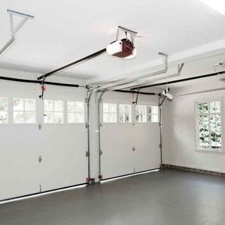 Empty white garage with a closed sectional door, gray floor, windows, and a ceiling-mounted opener