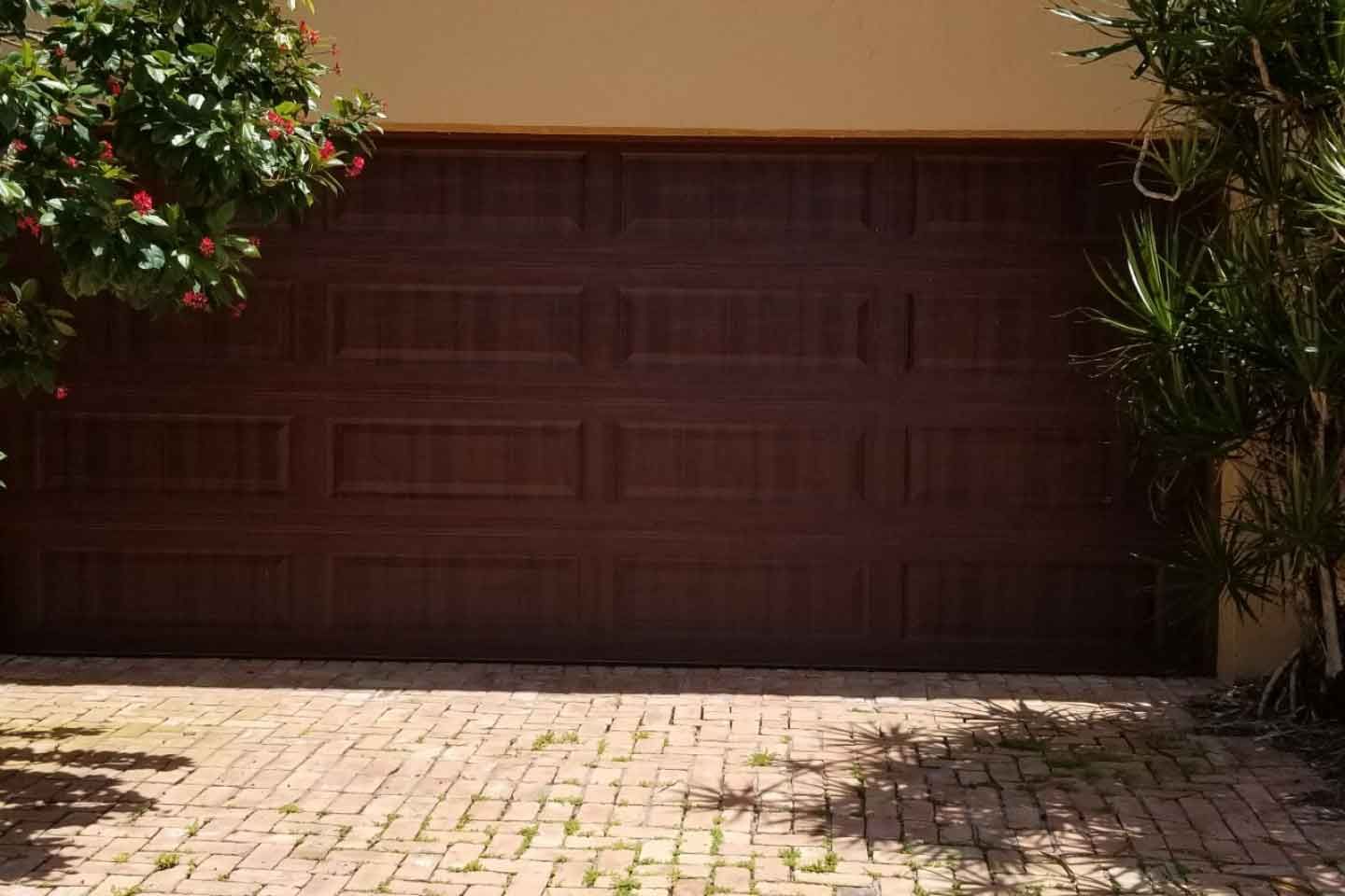 Closed dark brown garage door beneath beige wall, with sunlit brick driveway and plants at the sides