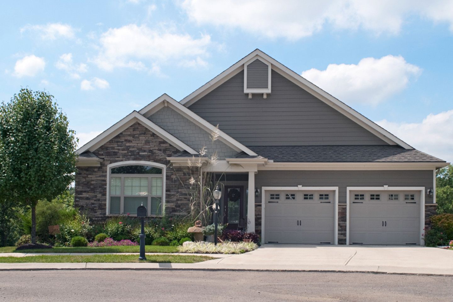 Suburban house with gray siding, stone accents, and a two-car garage under a blue sky.