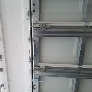 Close-up of a gray metal garage door track and hinge assembly against a white wall.