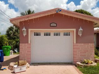 Pink garage with a white door, brick trim, and lanterns under a sunny sky