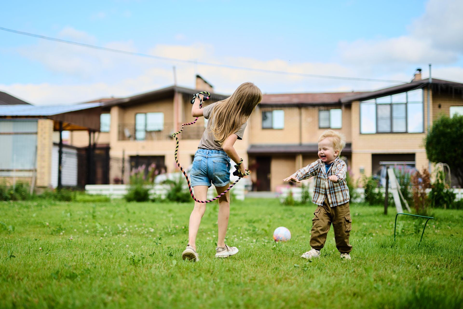 Two children play together on a grassy lawn in front of a large house