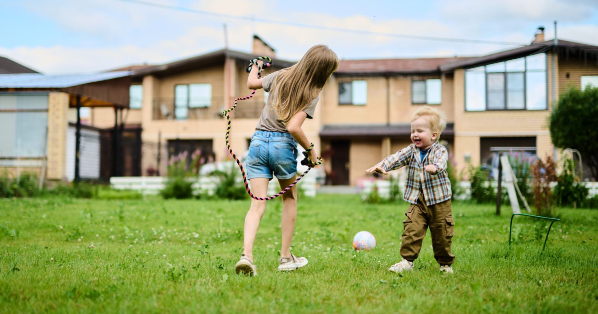 Two children play together on a lawn in front of a large house