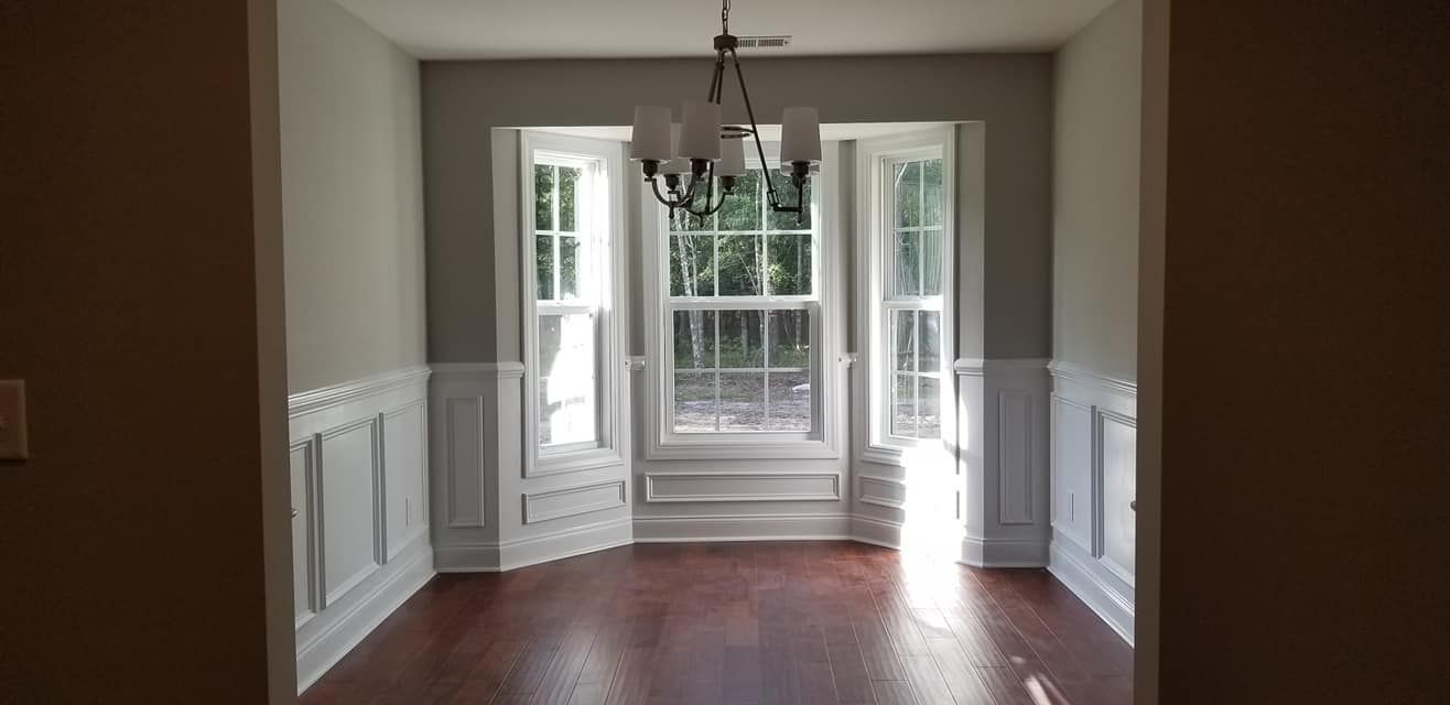 A bright dining room with hardwood floors, white wainscoting, light gray walls, a bay window, and a hanging chandelier.
