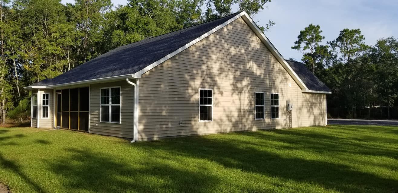 A light-colored, single-story house with a gabled roof and screened porch, set against a backdrop of green trees.