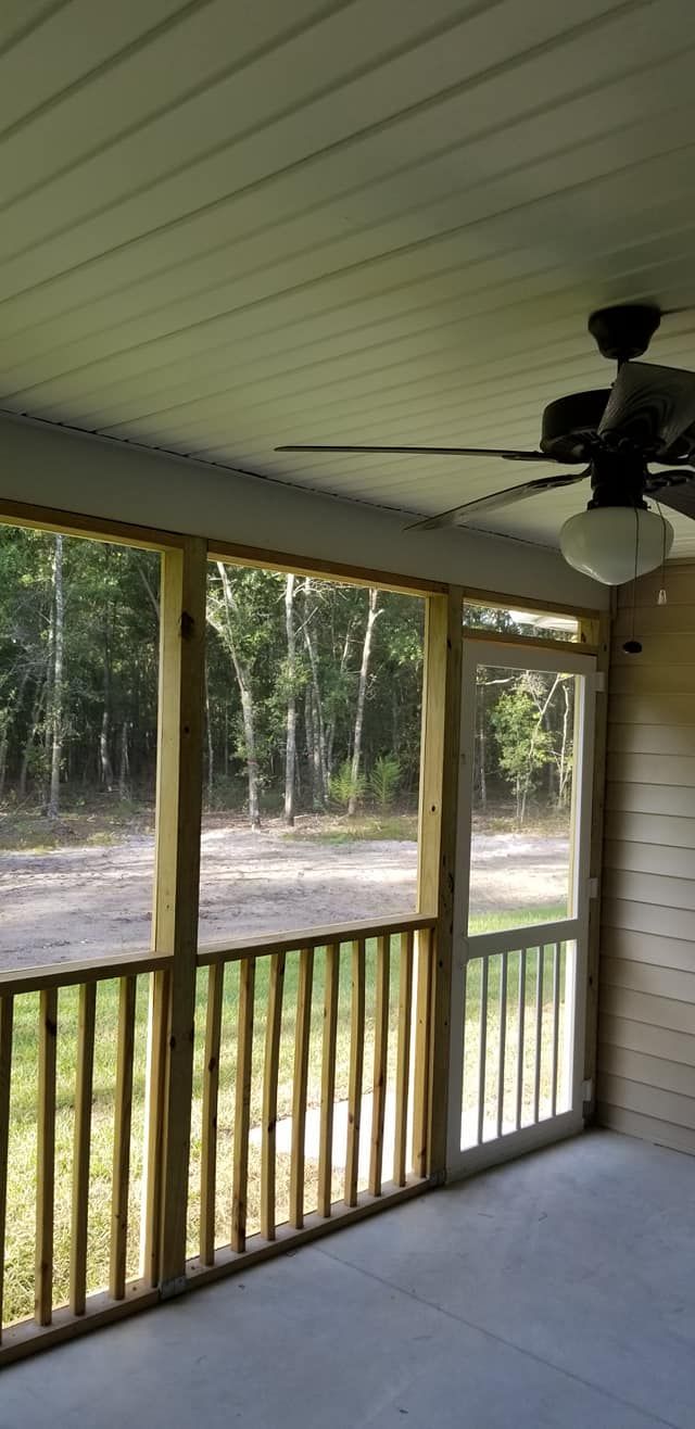 A screened-in porch with wood railing, concrete floor, white ceiling, ceiling fan, and a door looking out onto a woods.