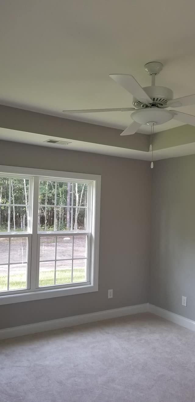 A room with neutral gray walls, a white ceiling fan, a window looking outside, and light-colored carpeting.