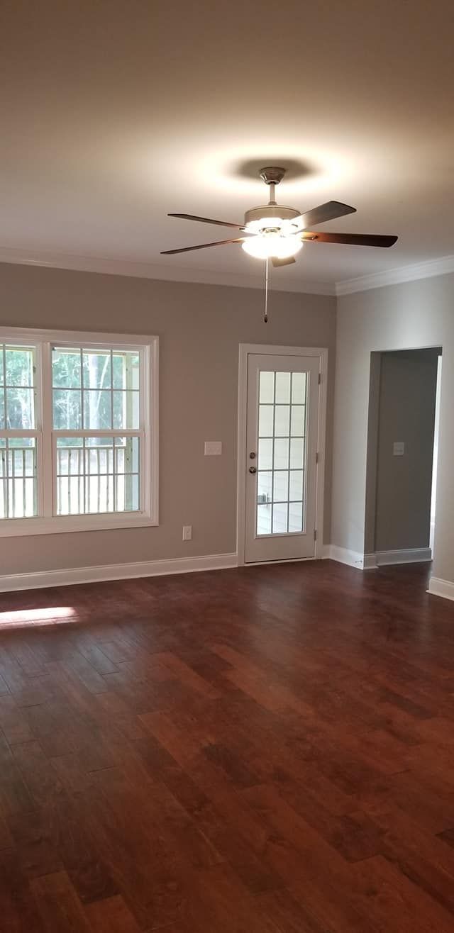 A bright, empty room with hardwood floors, a ceiling fan, a window looking outside, a glass-pane door, and a doorway.