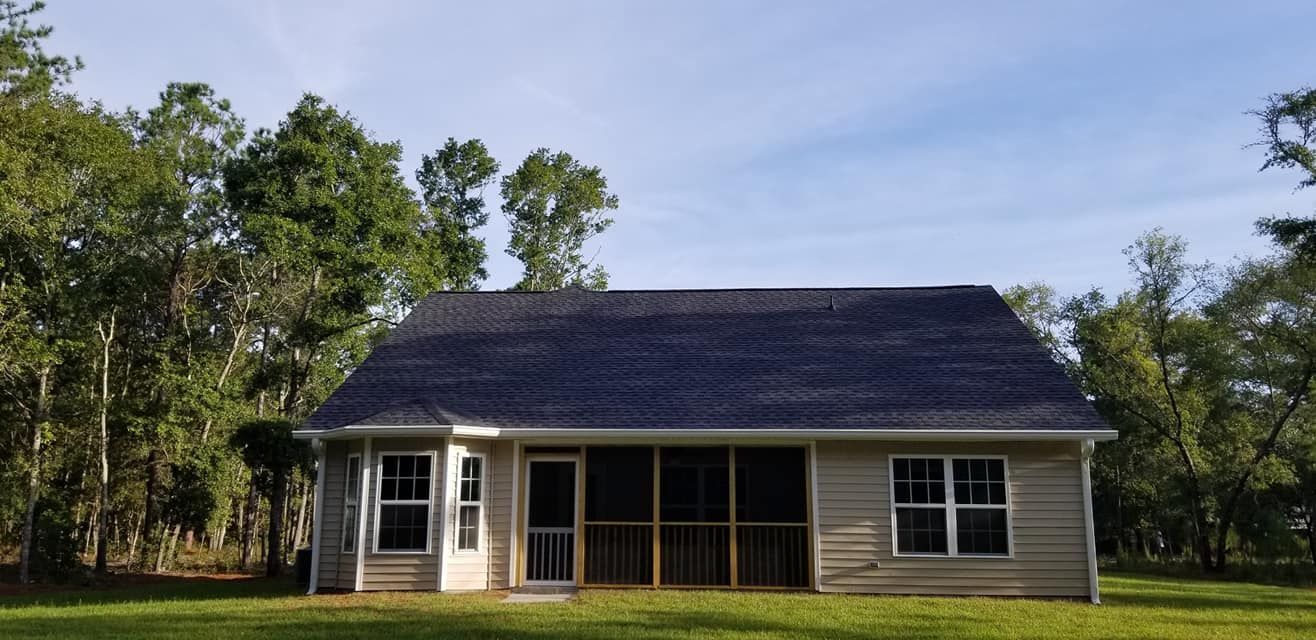 A one-story tan house with a dark roof and a central screened porch, surrounded by trees on a sunny day.