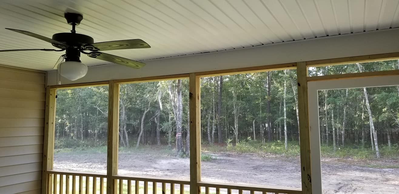 A screened-in porch with a ceiling fan, light fixture, and view of a wooded area outside.