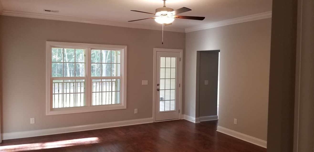 An empty room with light tan walls, hardwood floors, a ceiling fan, a large window with white trim, and a glass door.