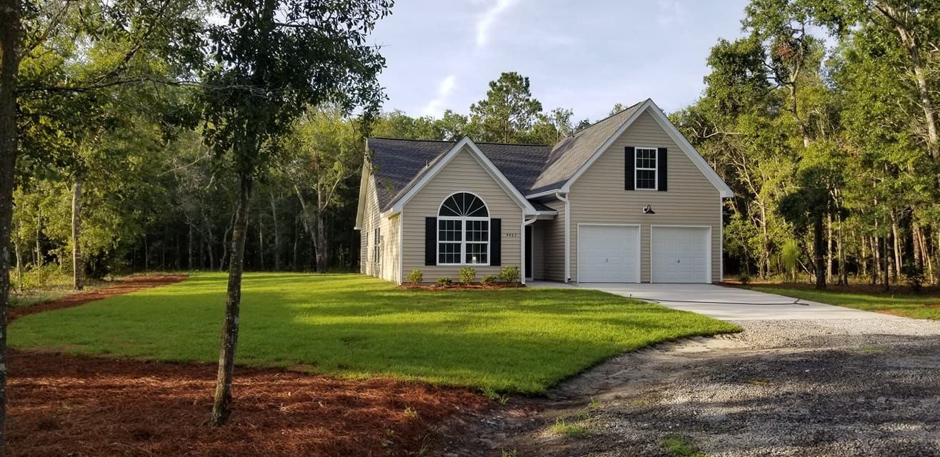 A beige, two-story house with a two-car garage, surrounded by green lawns and dense woods under a bright sky.