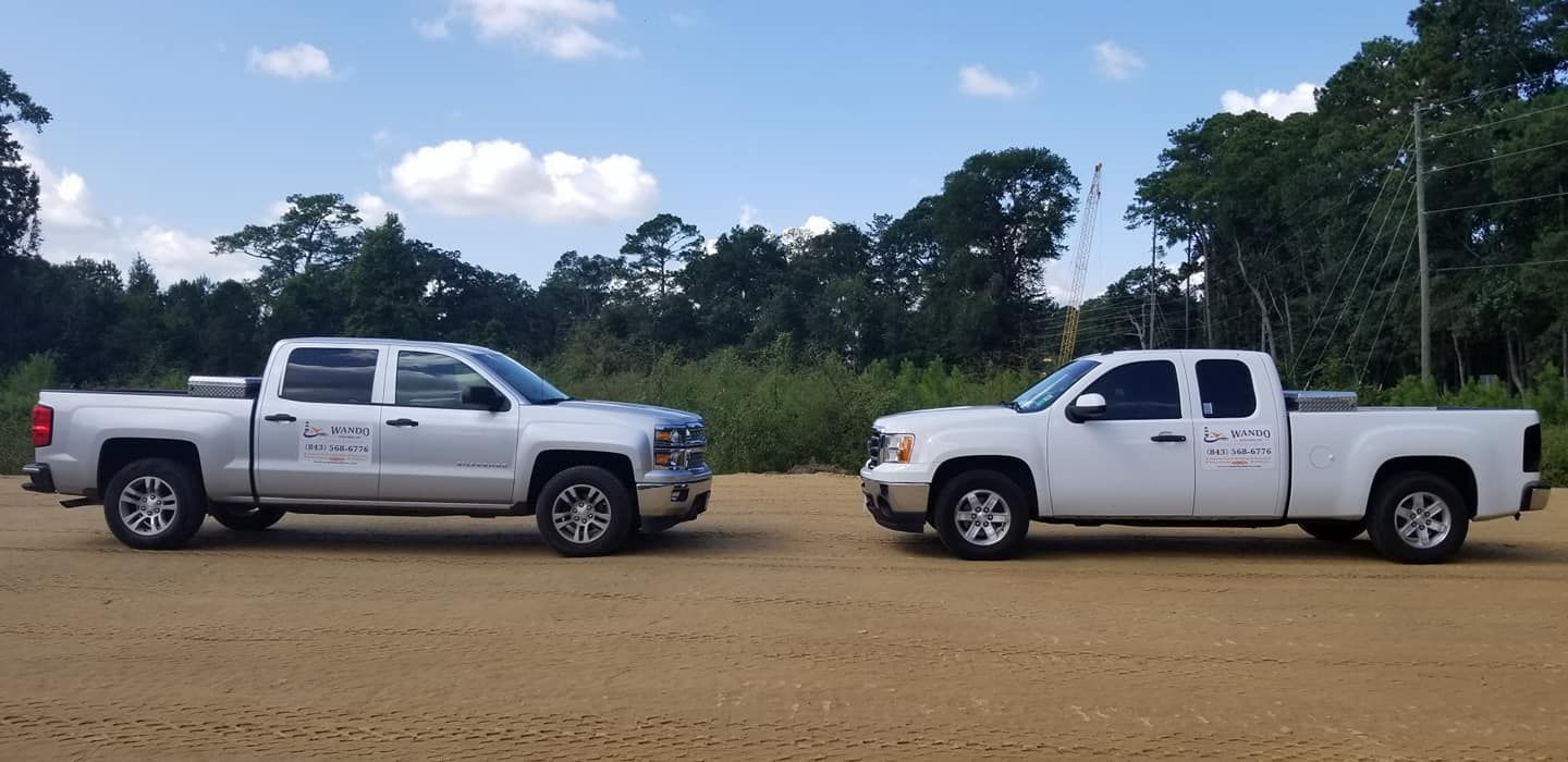 Two pickup trucks, one silver and one white, parked side-by-side on a dirt lot with a treeline in the background.