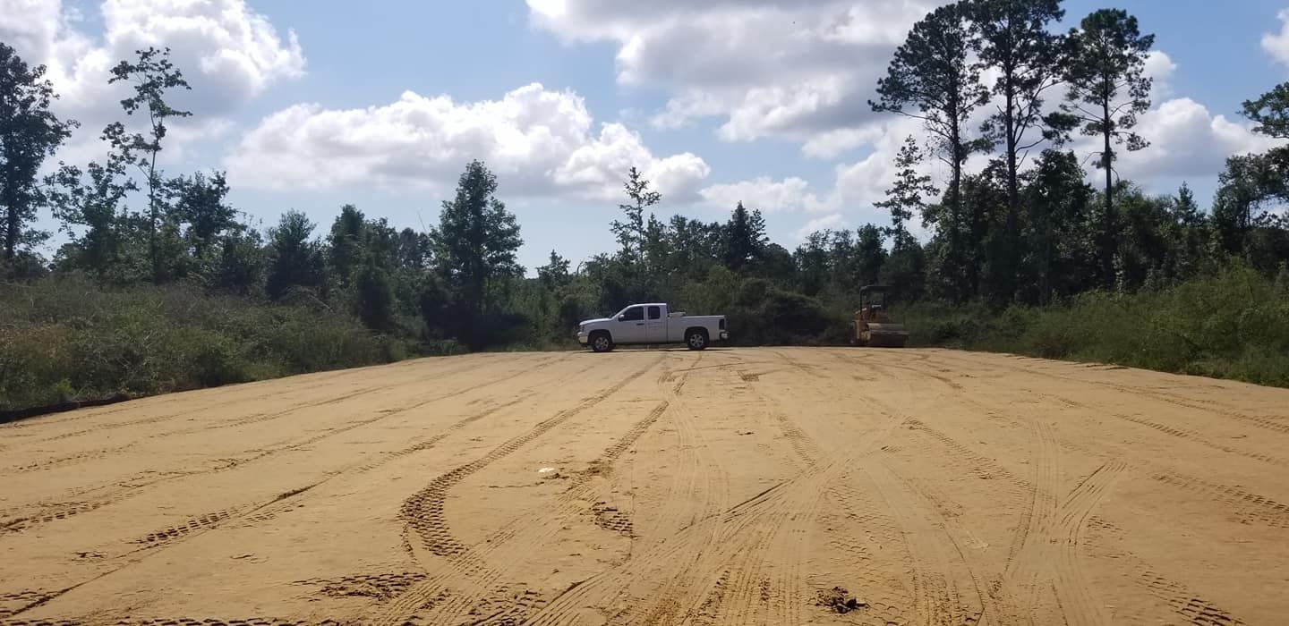 A white pickup truck parked on a leveled, sandy lot surrounded by a wooded area under a blue, cloudy sky.