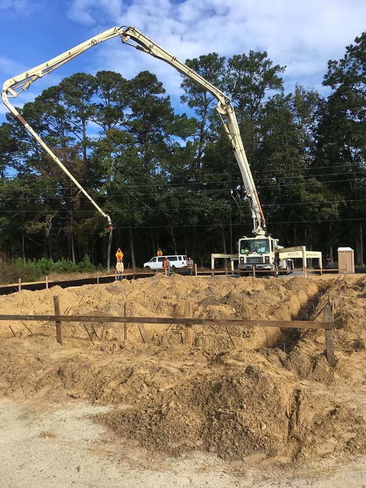 A white concrete pump truck extends its long boom arm over a dirt construction site with wooden framing in the foreground.