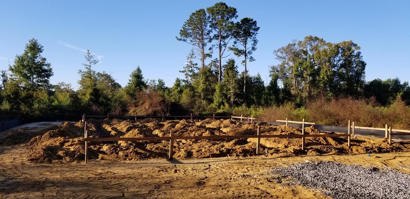 A construction site with wooden stakes and string marking a foundation, surrounded by dirt, trees, and a clear blue sky.