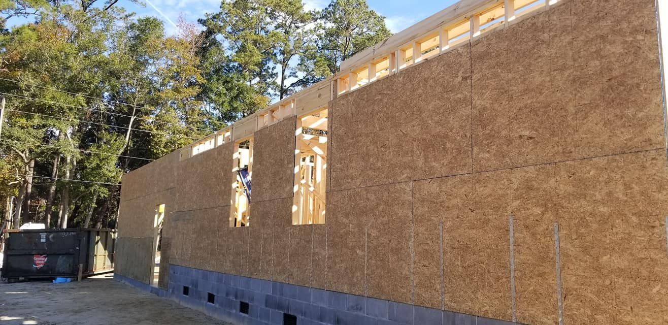 A partially constructed building frame with OSB sheathing walls, sitting on a gray block foundation near a forest.