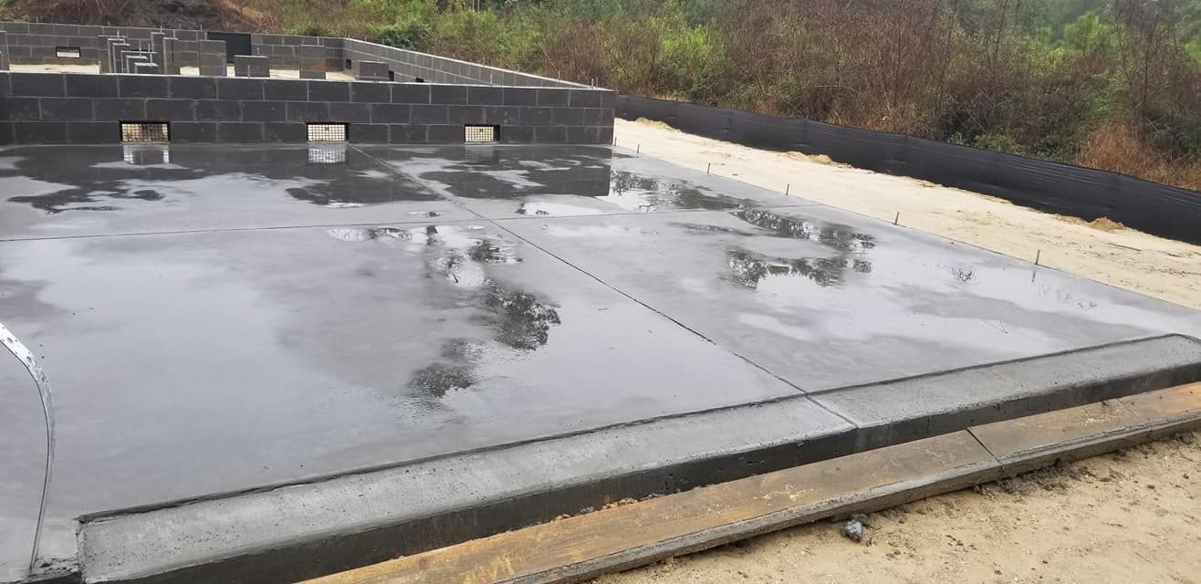 A freshly poured concrete foundation slab with cinder block walls, surrounded by dirt and vegetation.