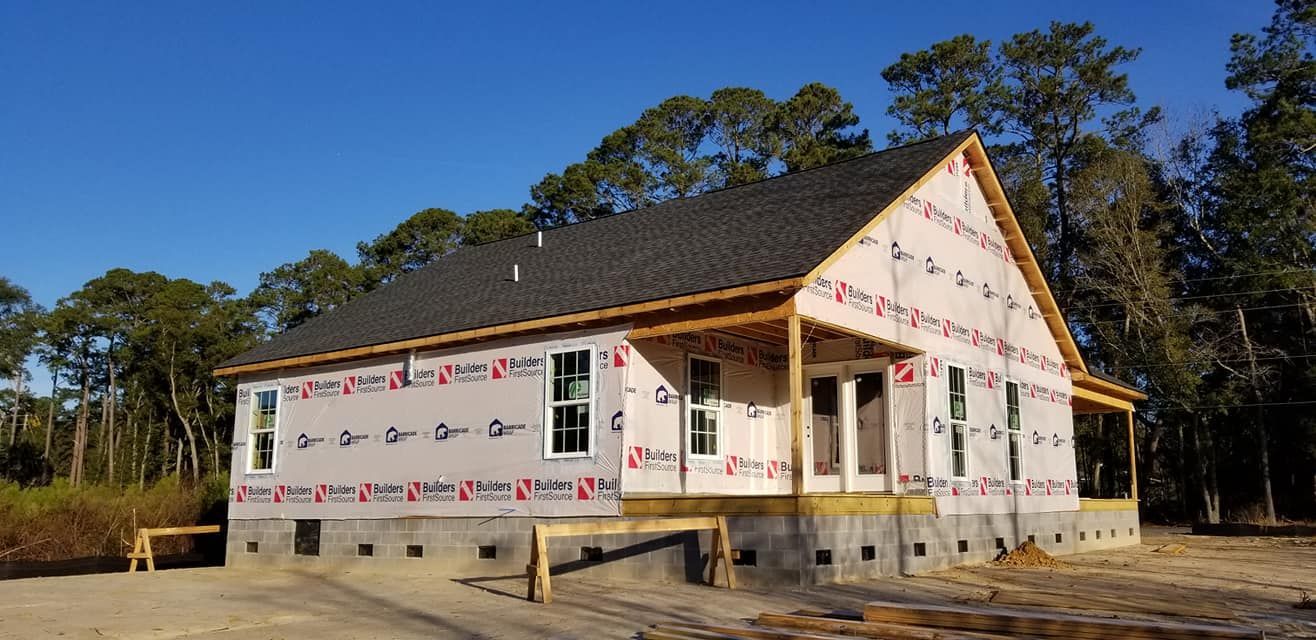 A house under construction with a gray roof and protective building wrap on the exterior walls, set against a forest.