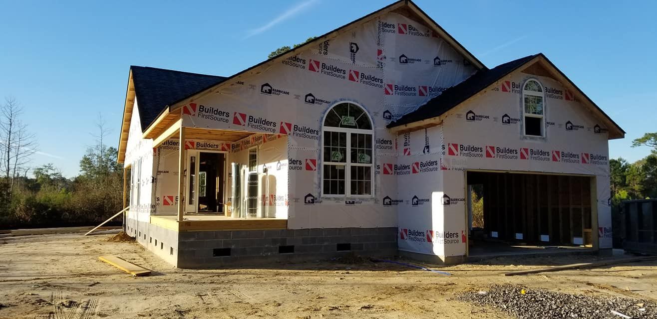 A house under construction with exposed wooden framing and white weather-resistant wrap on a dirt lot.