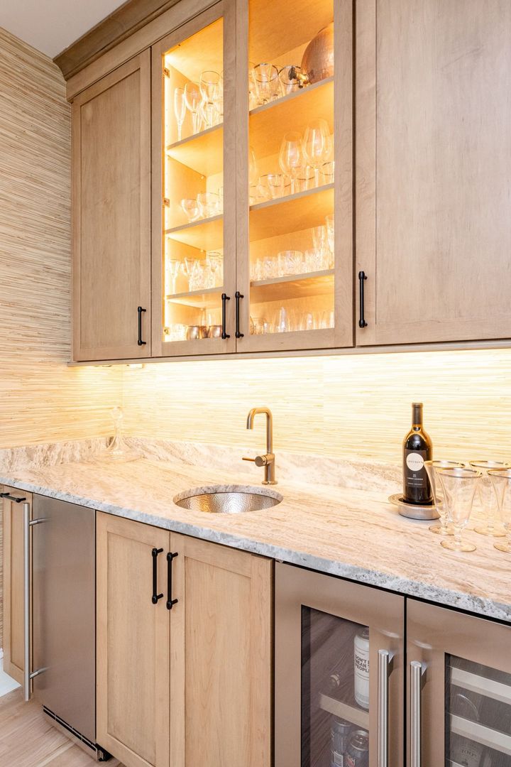 A modern home bar with light wood cabinetry, illuminated glass cabinets, a granite countertop, a sink, and a wine fridge.