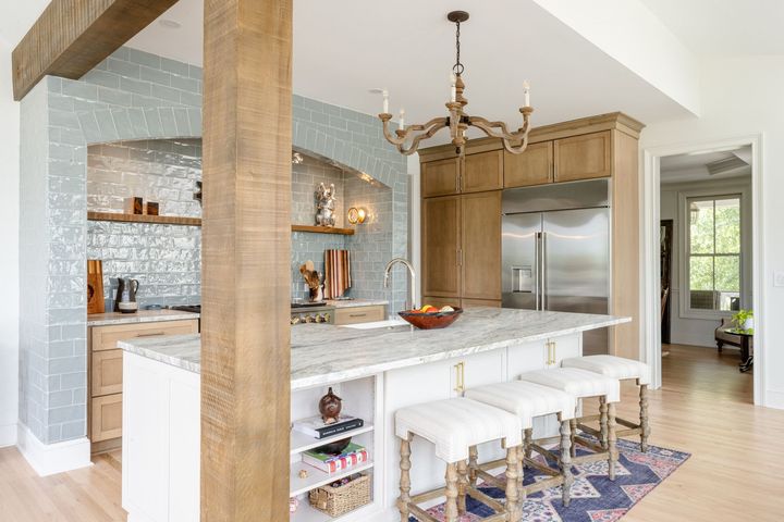 A rustic-modern kitchen with light wood cabinets, a marble island, white bar stools, and a pale blue tile backsplash.