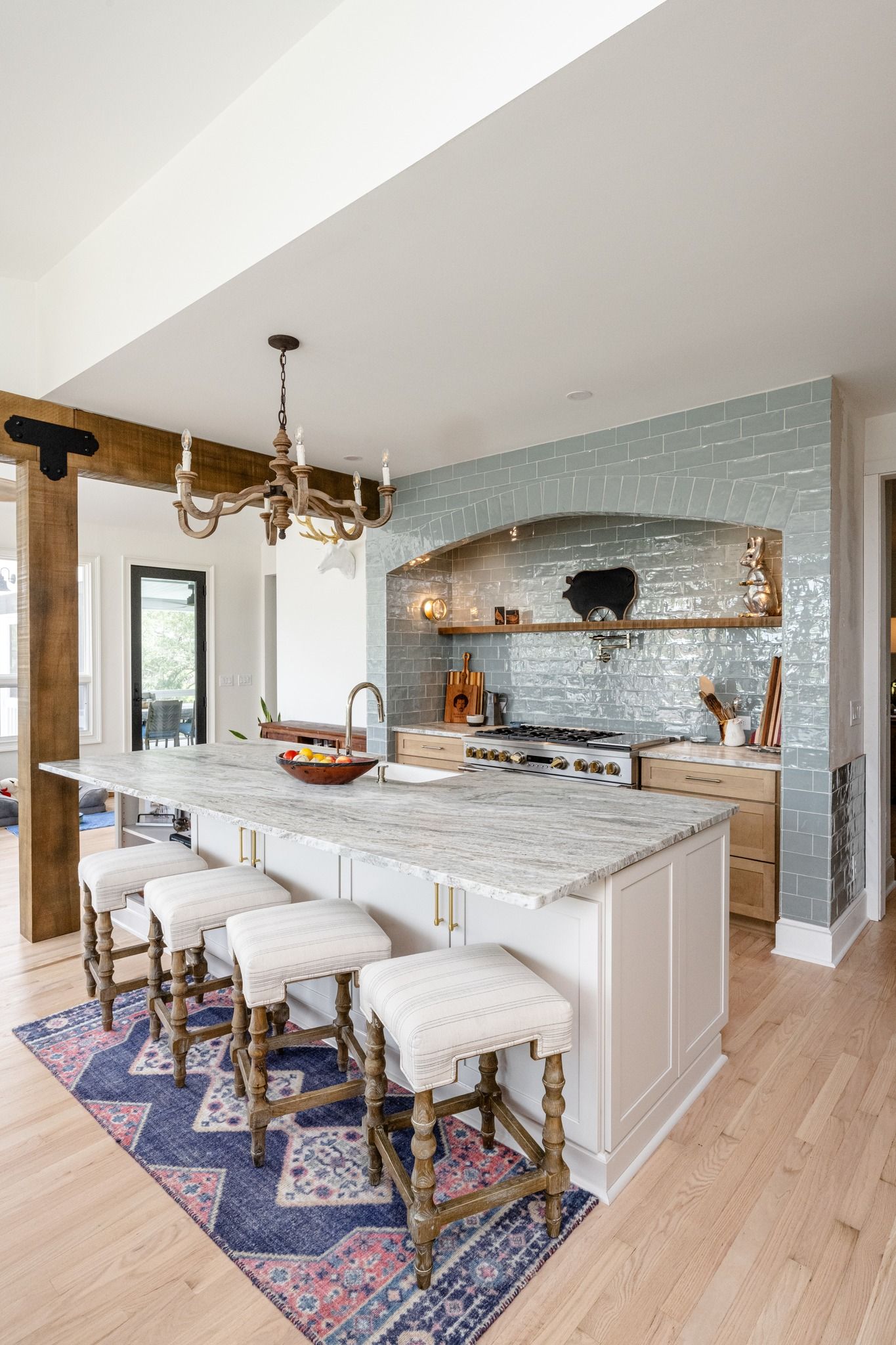 A bright kitchen with a marble-topped island, wooden stools, a colorful runner rug, and a tiled wall above the stove.