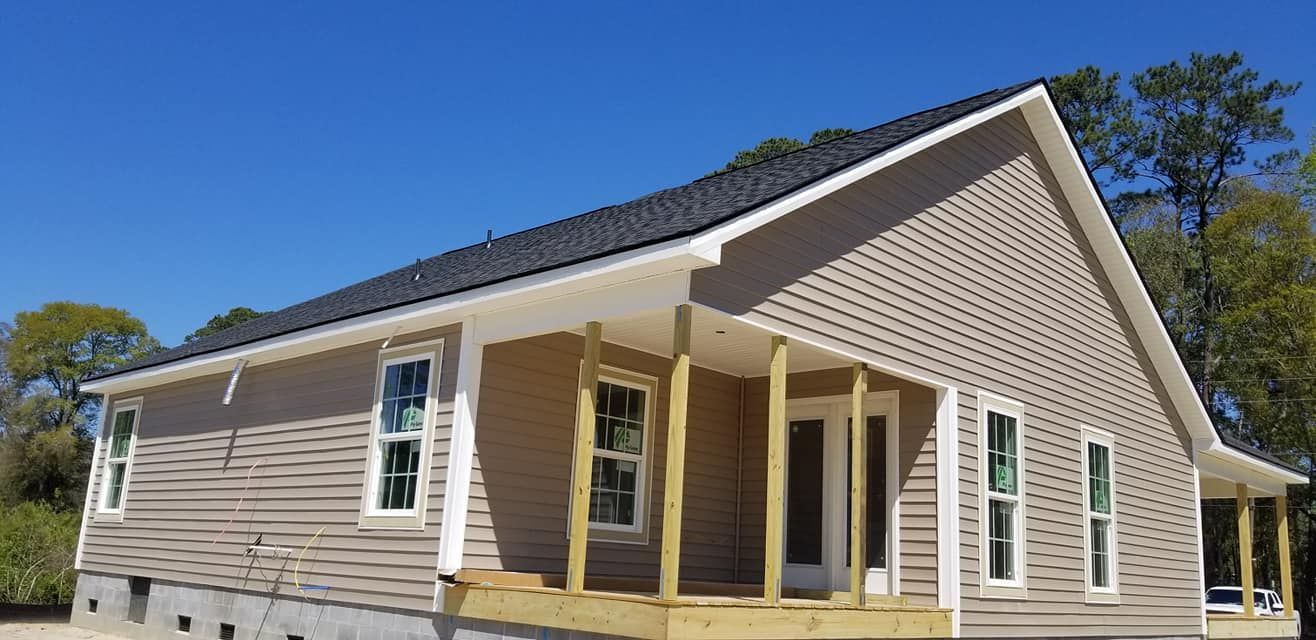 A tan, single-story house with a black roof and an unfinished wooden front porch, surrounded by trees under a blue sky.