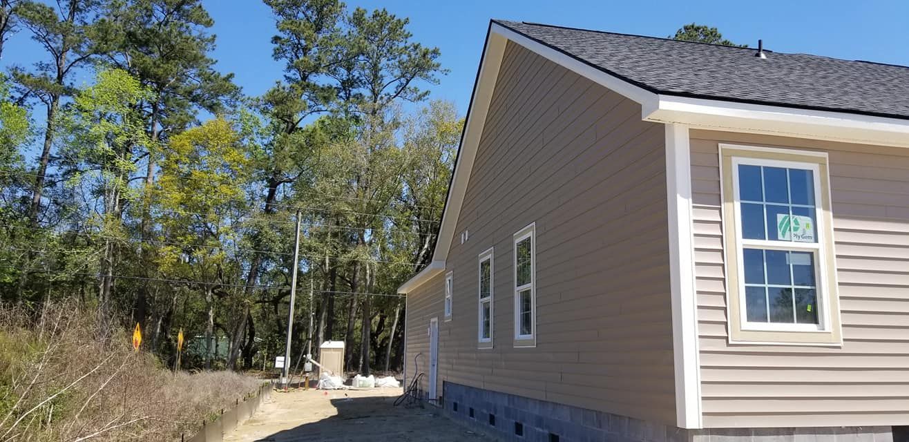 Tan-sided house under construction with a dark shingled roof, next to a wooded area on a sunny day.