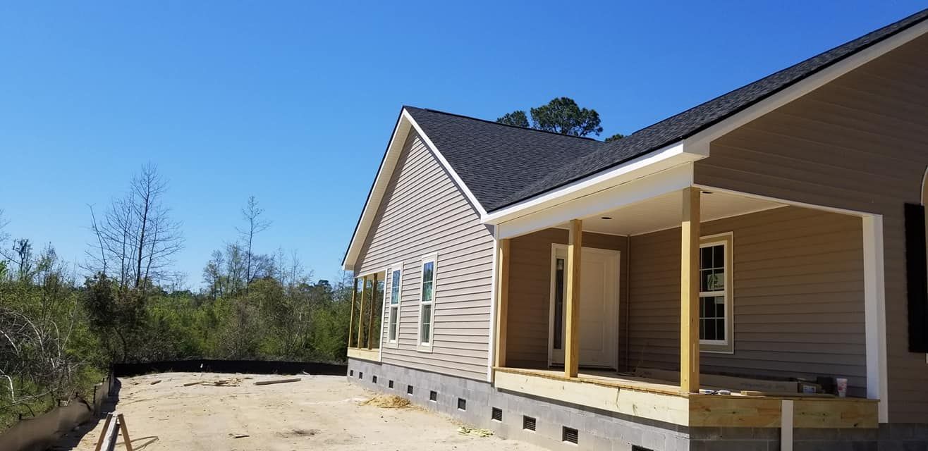 A newly constructed house with tan siding and a dark roof, featuring a front porch, on a sandy lot with trees.