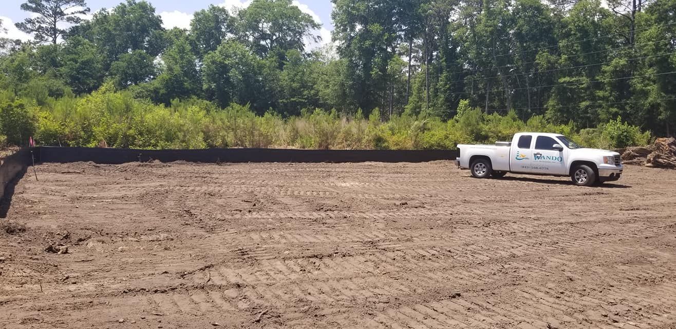 A white pickup truck parked on a cleared, level plot of dirt next to a dense line of trees on a sunny day.