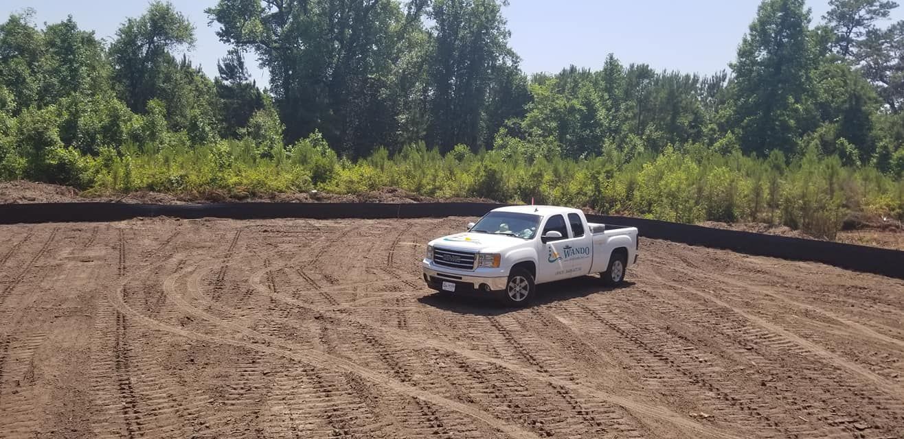 A white pickup truck parked in the center of a cleared dirt construction site bordered by trees.