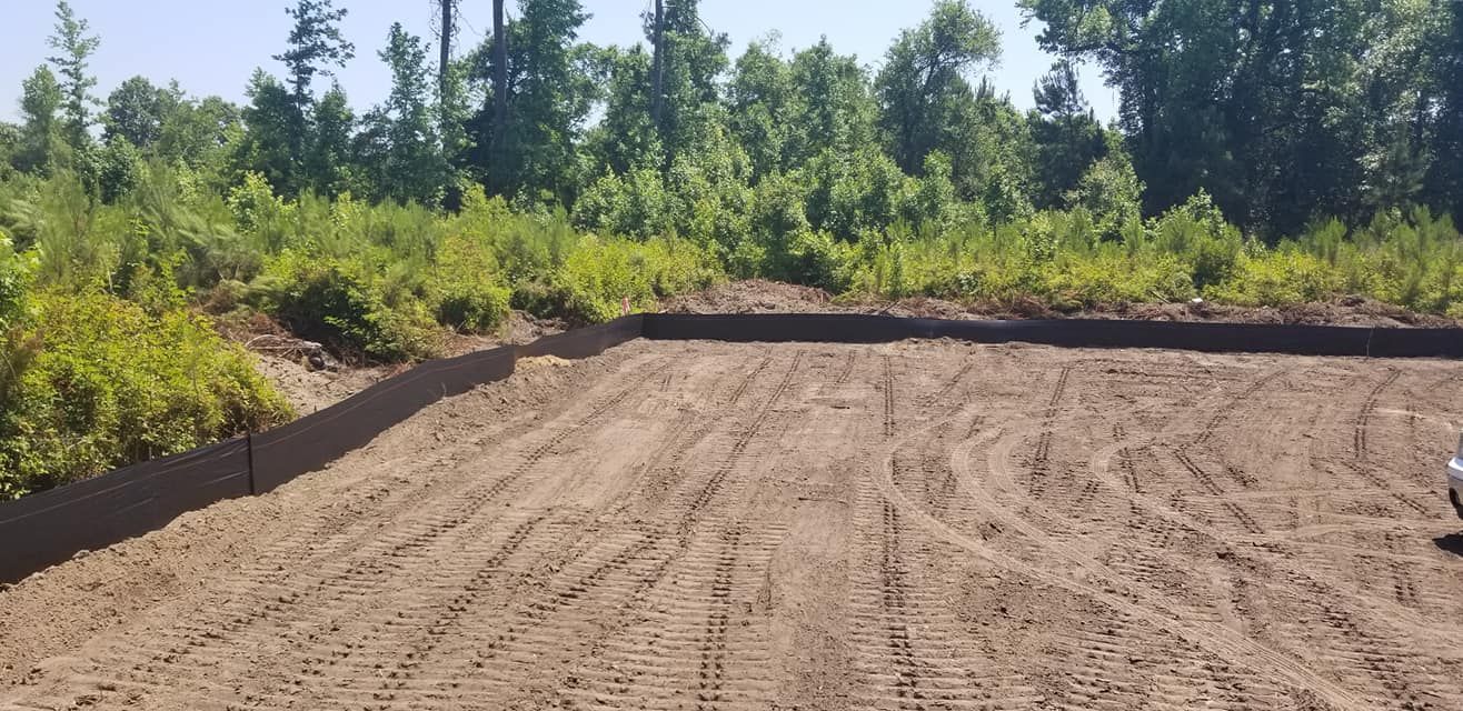A cleared dirt construction site surrounded by trees, with a black silt fence along the edge of the graded land.