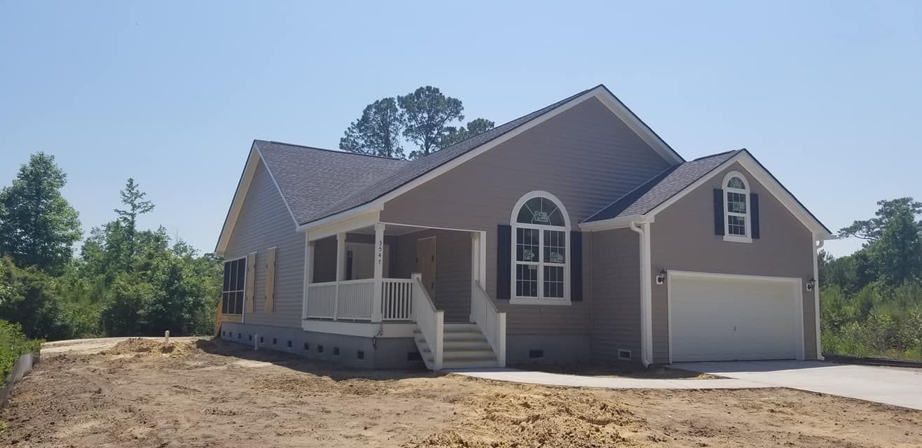 A newly constructed, gray-sided single-story house with a white front porch and attached garage on a cleared lot.