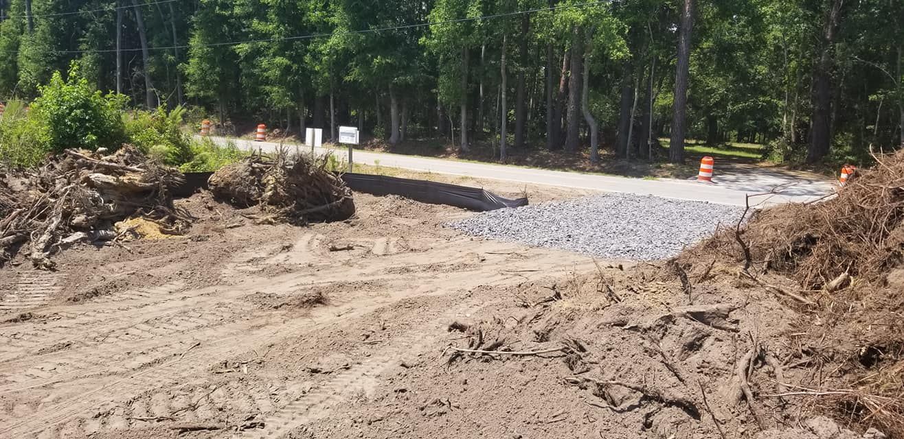 A gravel driveway construction site meets a paved road, surrounded by dirt piles and a dense forest background.