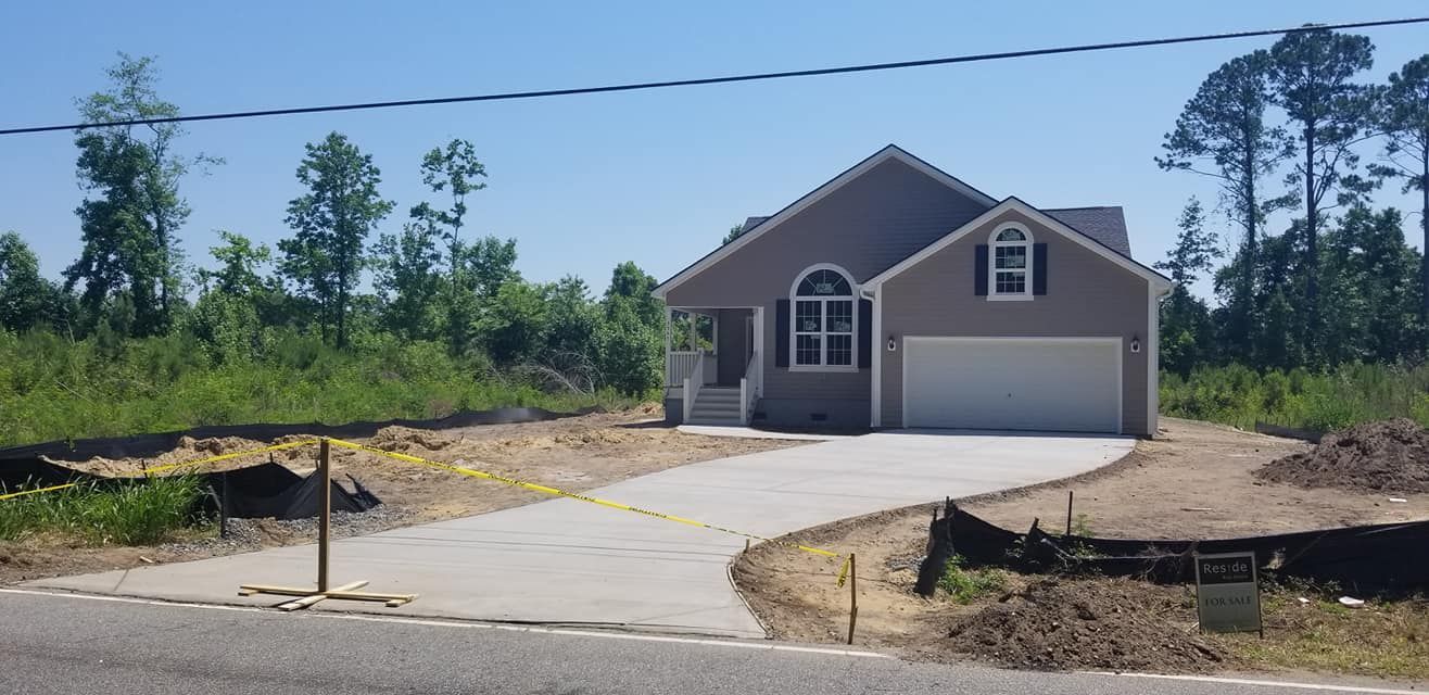 A newly constructed, single-story gray house with a white garage and curved concrete driveway in a wooded area.