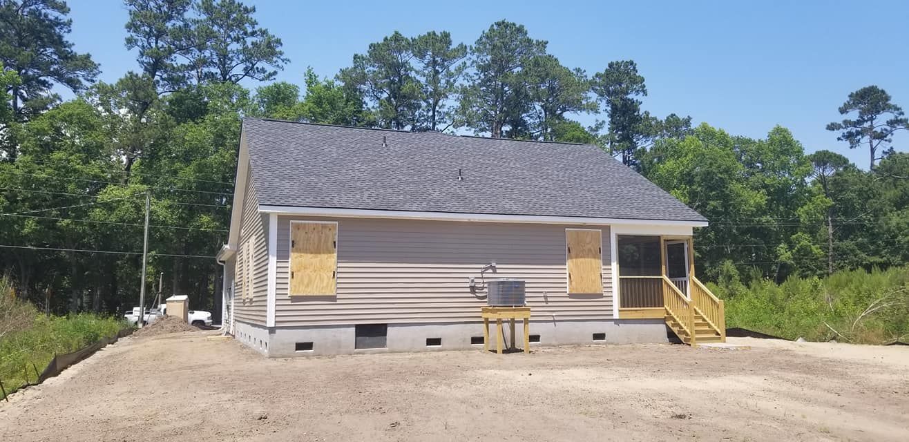 A newly constructed, single-story house with gray siding, a dark shingled roof, and boarded-up windows on a dirt lot.