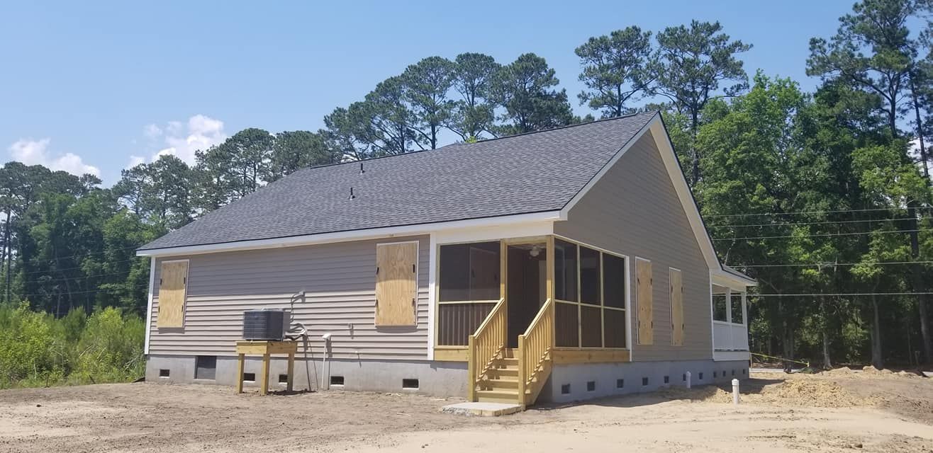 A beige house with a grey roof, boarded windows, a screened porch, and an outdoor HVAC unit on a wooden stand.
