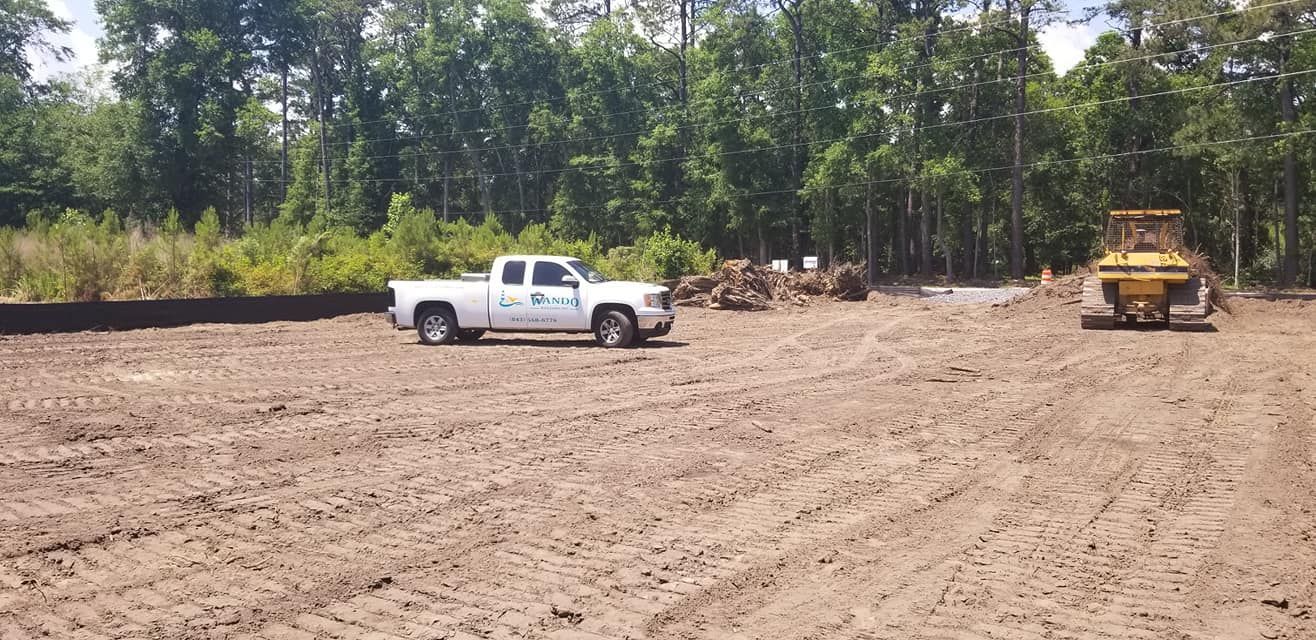 A white pickup truck and a yellow bulldozer parked on a clearing of leveled dirt in front of a dense forest.