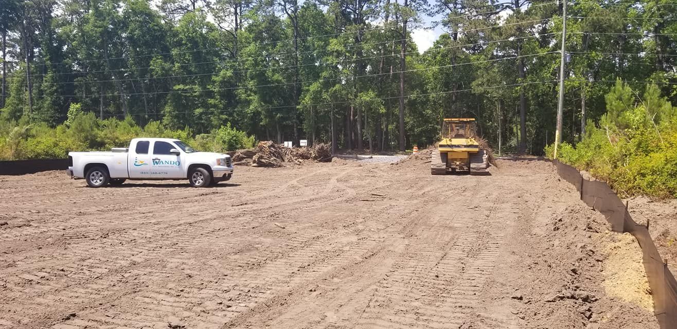 A construction site with a white pickup truck parked on dirt, a yellow bulldozer in the distance, and a silt fence.