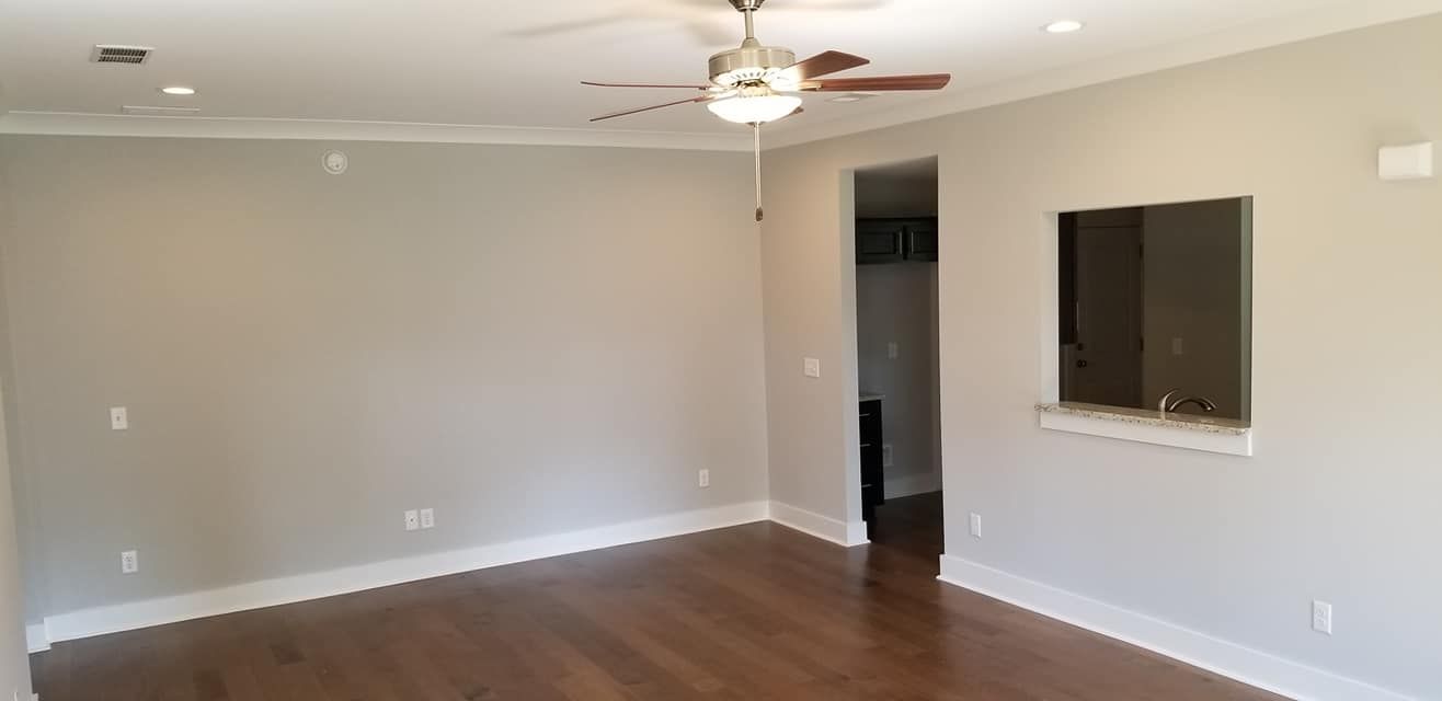 An empty room with light gray walls, dark wood floors, a ceiling fan, a doorway, and a kitchen pass-through window.