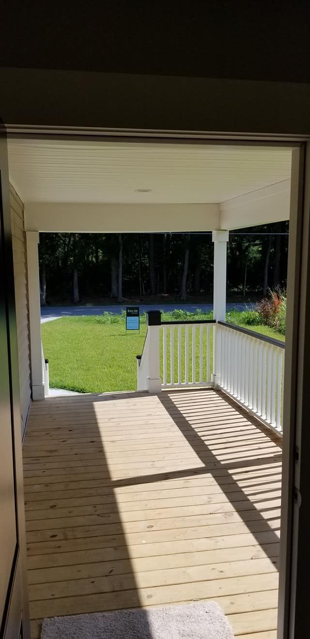 View from a doorway onto a wooden front porch with white railings, looking out over a grassy yard and trees.